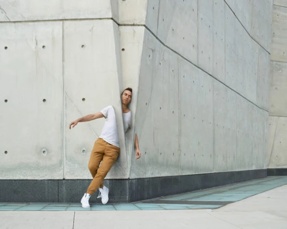 Adult male dancer in a white tee and tan pants leans against an angled concrete wall outdoors in an urban setting, captured in medium shot.