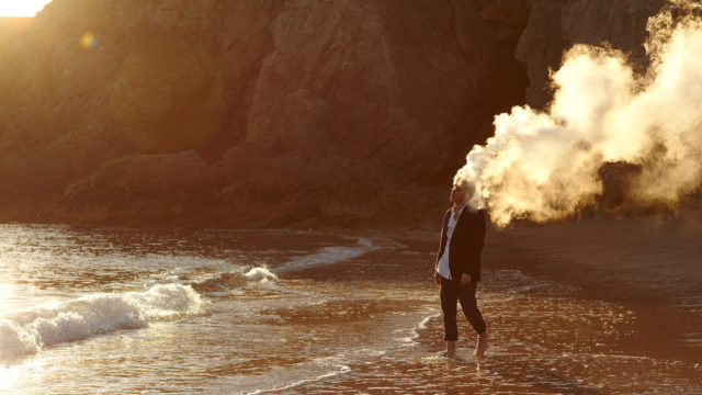 Man stands on a beach with a large cloud of smoke emanating from his head, ocean waves, sand, and coastal cliffs.