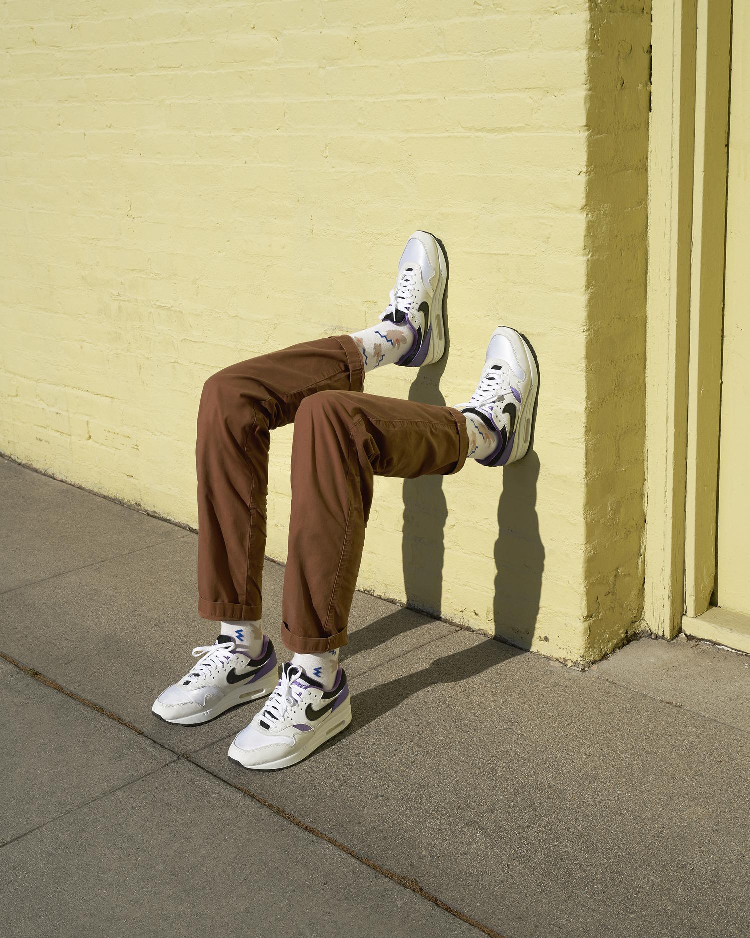 Legs wearing brown trousers and white sneakers appear to emerge from a light yellow building, positioned on a grey sidewalk in an outdoor setting.