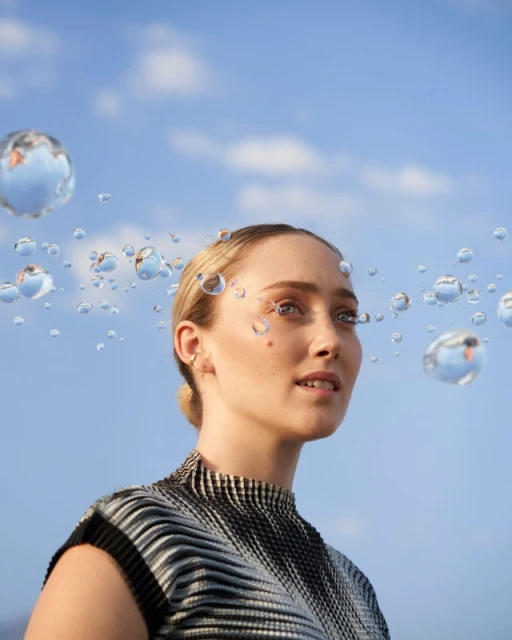 Woman with blonde hair styled back in an updo, wearing a black and white striped shirt, posing in front of a blue sky with clouds.