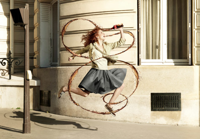 Red-haired woman in a grey skirt and tan jacket jumps, drinks a soda with brown liquid swirls around her in front of an urban building.