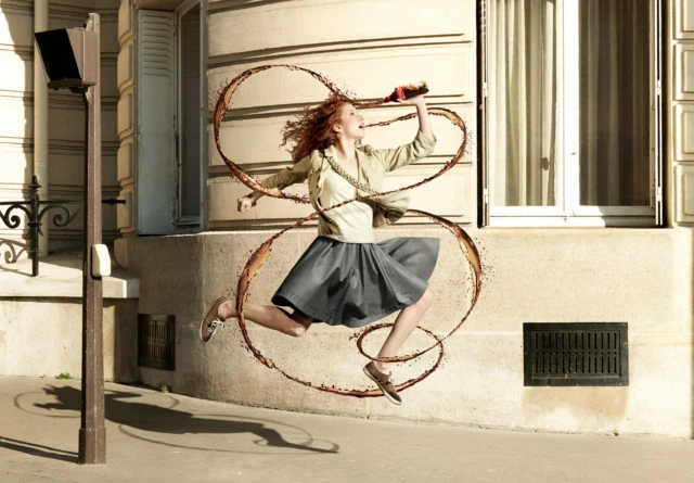 Red-haired woman in a grey skirt and tan jacket jumps, drinks a soda with brown liquid swirls around her in front of an urban building.