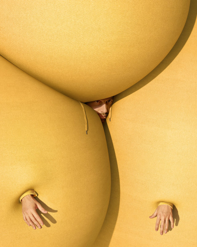 Surreal studio portrait featuring a person peering from between oversized yellow fabric forms, with hands emerging from openings.