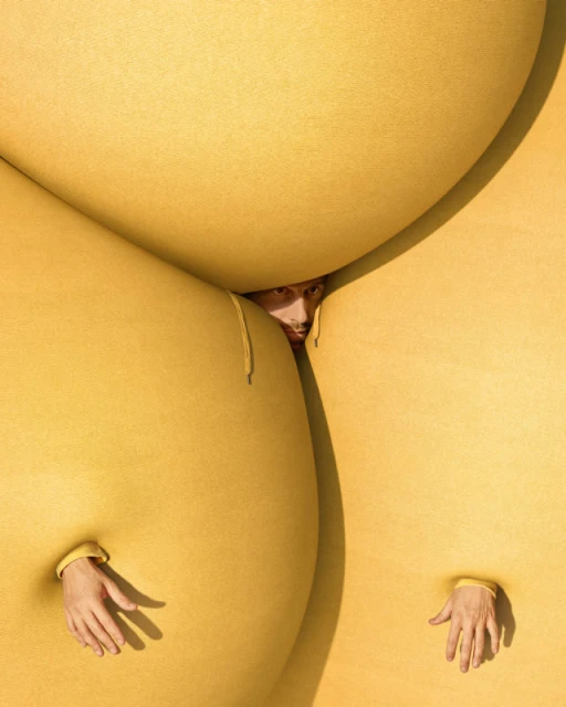 Surreal studio portrait featuring a person peering from between oversized yellow fabric forms, with hands emerging from openings.