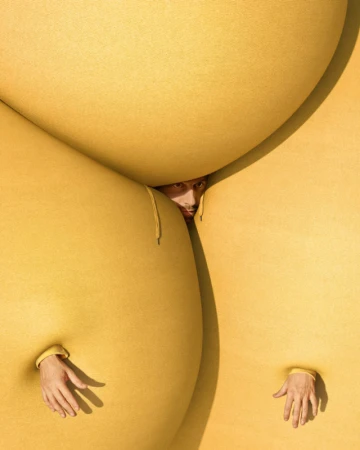 Surreal studio portrait featuring a person peering from between oversized yellow fabric forms, with hands emerging from openings.