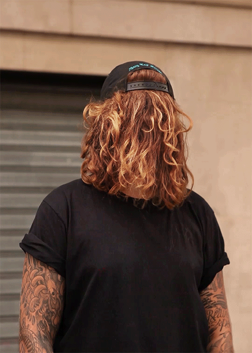Person in black T-shirt and baseball cap with long curly hair covering face standing against a building with neutral-colored stone blocks.