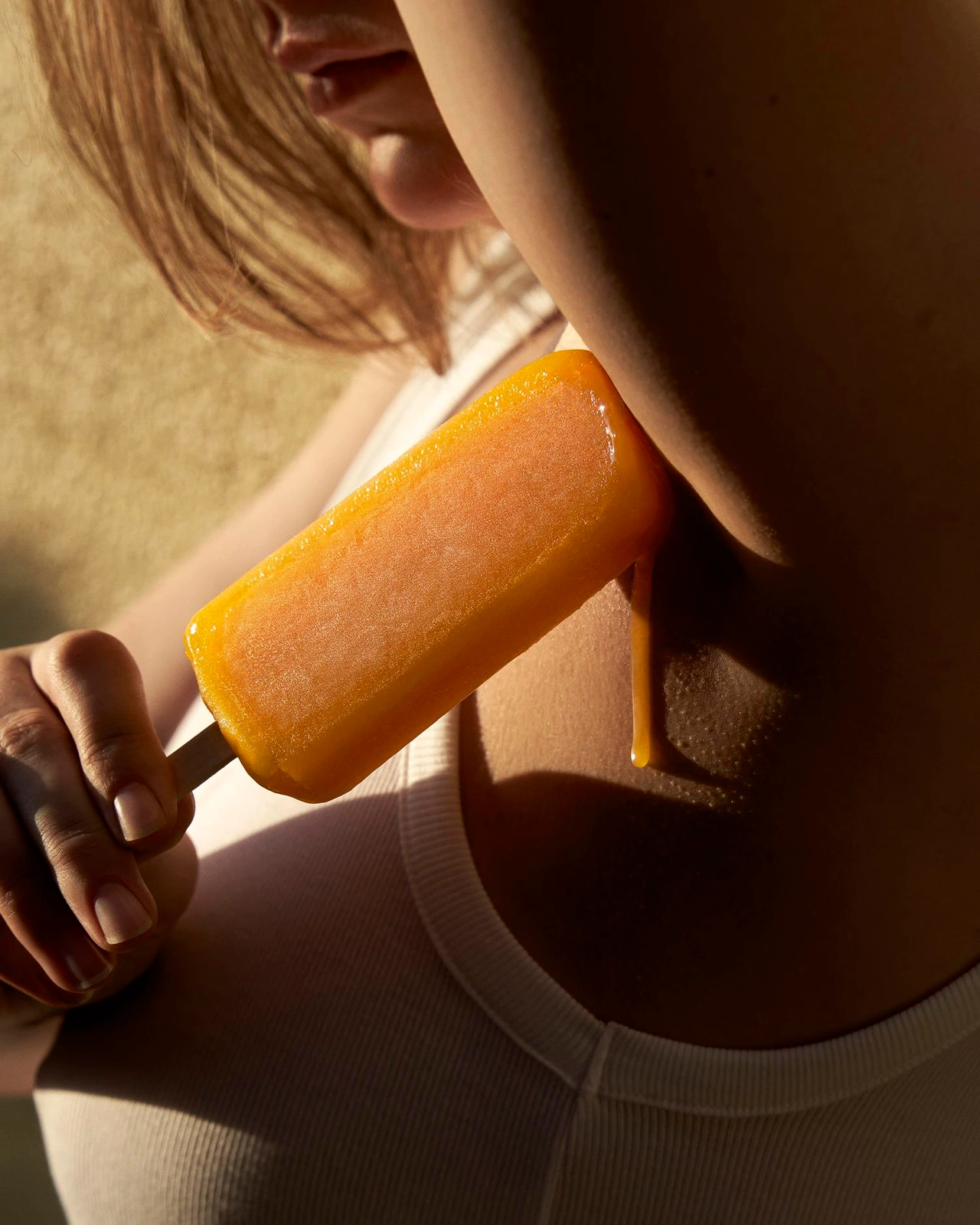 Close-up of a person holding a melting orange popsicle against their neck, wearing a white ribbed tank top.