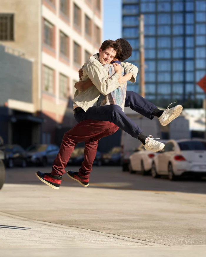 Two young men hugging mid-air outdoors on an urban street with parked cars and buildings in the background.