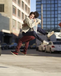 Two young men hugging mid-air outdoors on an urban street with parked cars and buildings in the background.