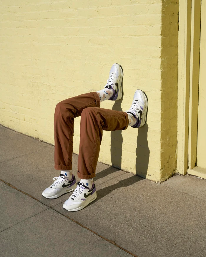 A set of legs wearing brown trousers and white sneakers pressed against a yellow brick wall outdoors.