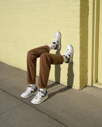 A set of legs wearing brown trousers and white sneakers pressed against a yellow brick wall outdoors.