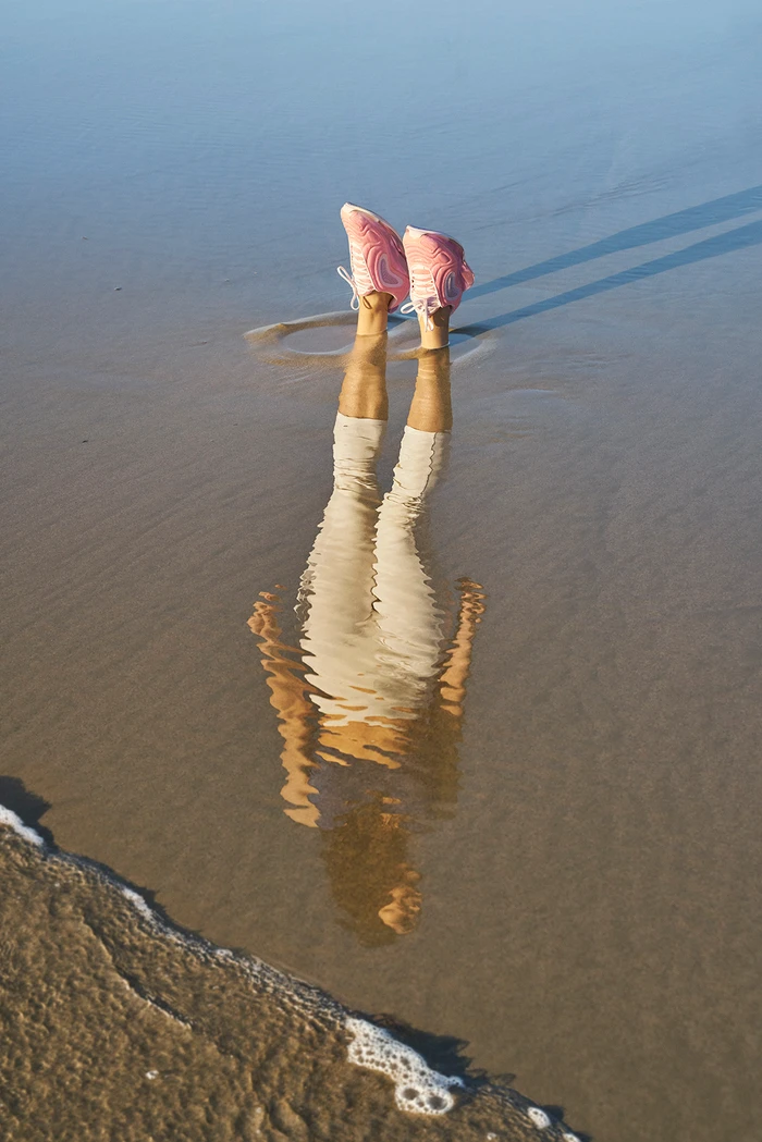 Legs in pink sneakers extending upward from a wet sandy beach with a rippled reflection on the water surface.