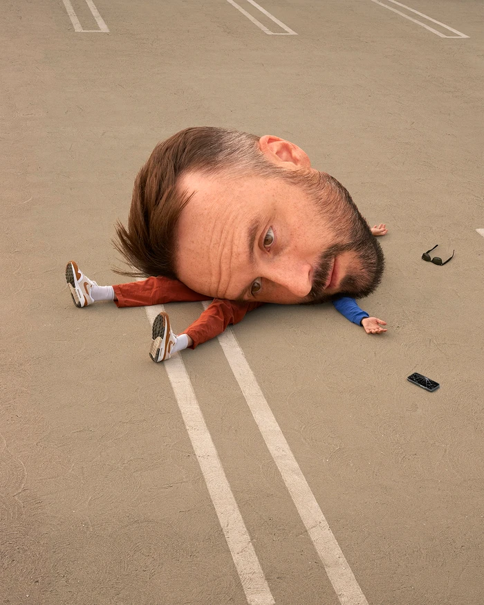 Surreal portrait by Romain Laurent featuring a man with an exaggerated giant head lying on an asphalt parking lot.
