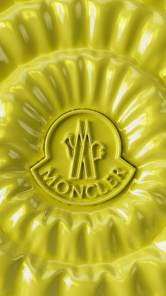 Close-up of a yellow Moncler logo on textured material. The logo features two stylized wings and the brand name in a circular emblem.
