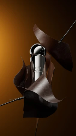 Metallic cylindrical perfume bottle with a chrome sphere cap surrounded by floating curved brown paper sheets against a warm background.