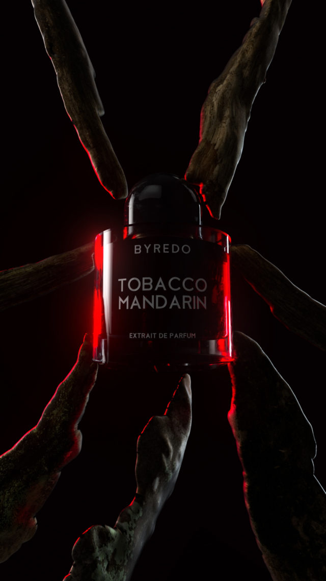 Dramatic product shot of Byredo Tobacco Mandarin perfume bottle surrounded by textured branches against a dark, moody background with red lighting.