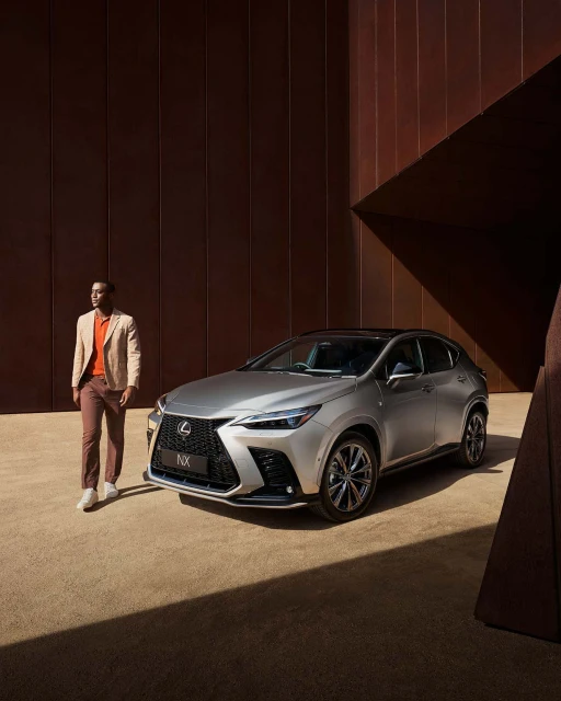 Silver Lexus NX parked on a tan surface with an adult male walking nearby and a textured copper wall behind, automotive setting.