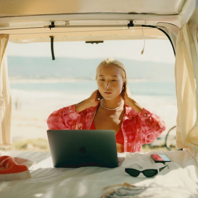 Young Asian woman using laptop inside camper van at a beach. She wears a red shirt. The background includes a sandy beach and ocean.