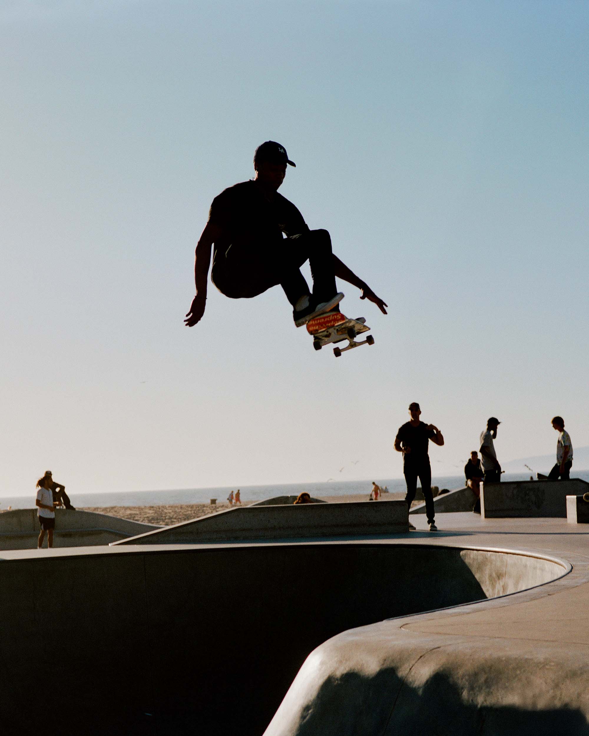 Silhouette of a skateboarder in mid-air at a skatepark with people and the ocean in the background on a sunny day with a clear sky.