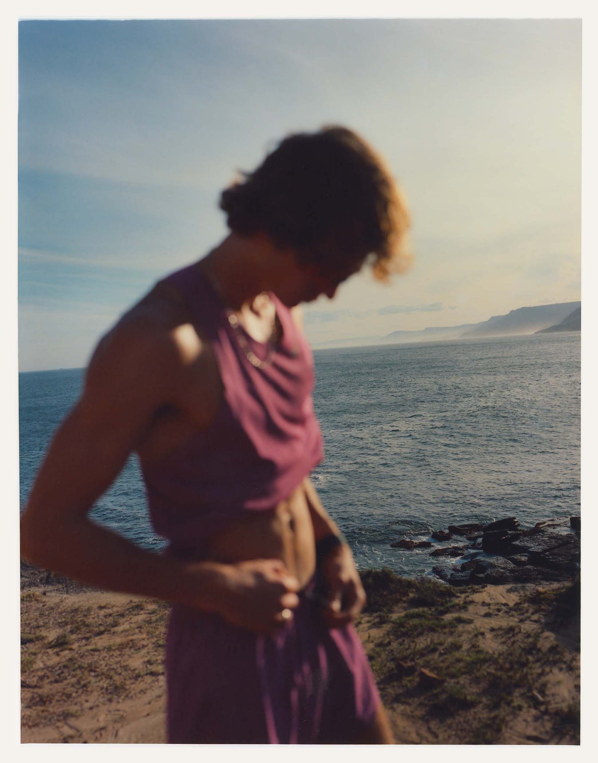 Blurred image of a young adult male on a beach, wearing a purple two-piece outfit, with the ocean and coastline visible in the background.