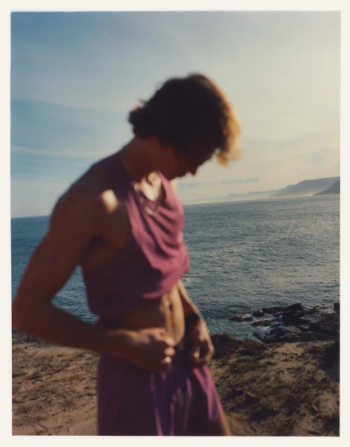 Blurred image of a young adult male on a beach, wearing a purple two-piece outfit, with the ocean and coastline visible in the background.