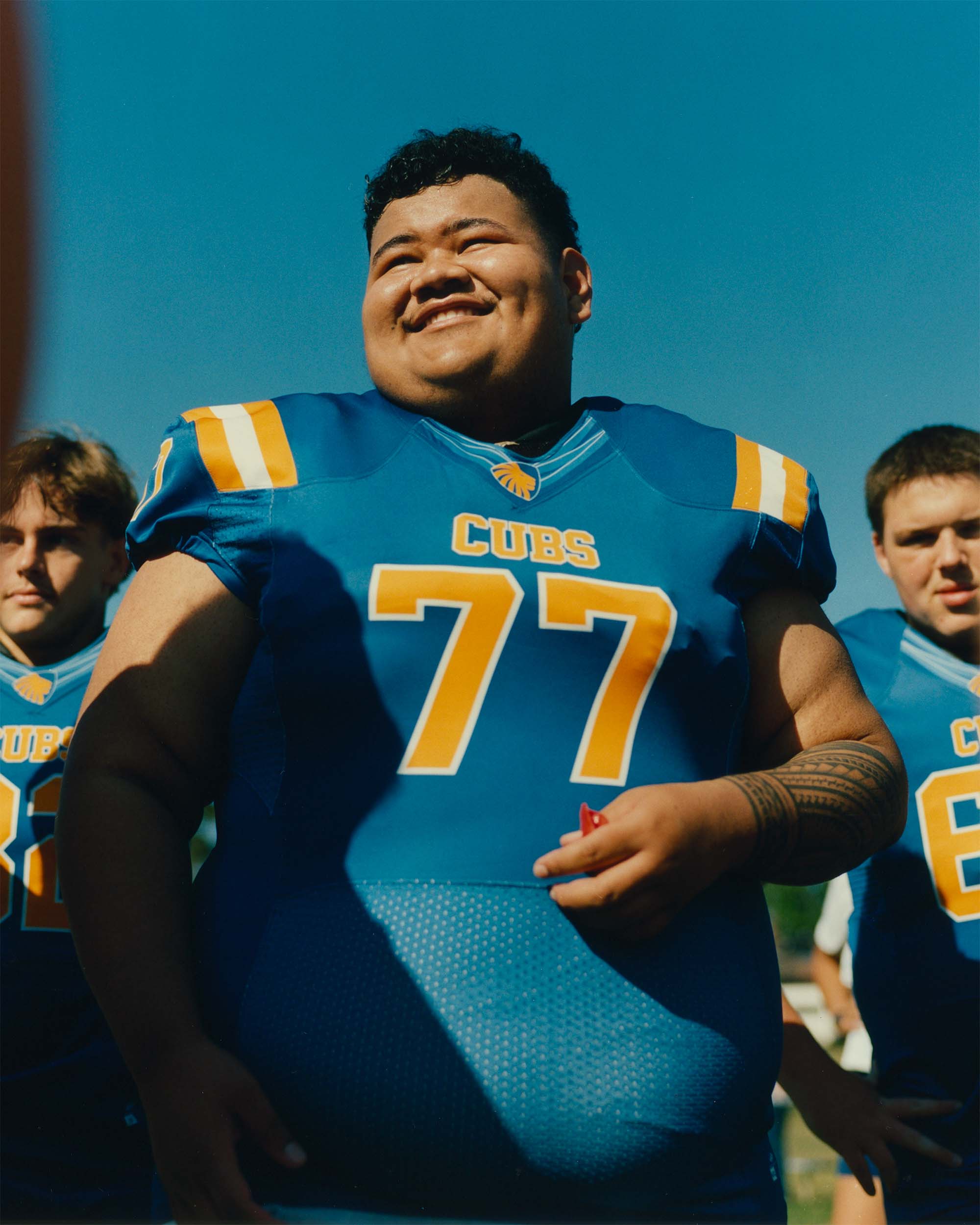 Football player in blue uniform smiles outdoors with teammates in the background on a sunny day, focus on central player.