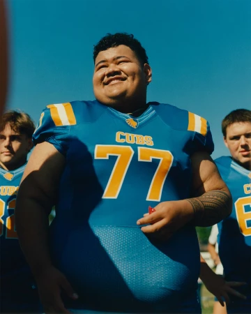 Football player in blue uniform smiles outdoors with teammates in the background on a sunny day, focus on central player.