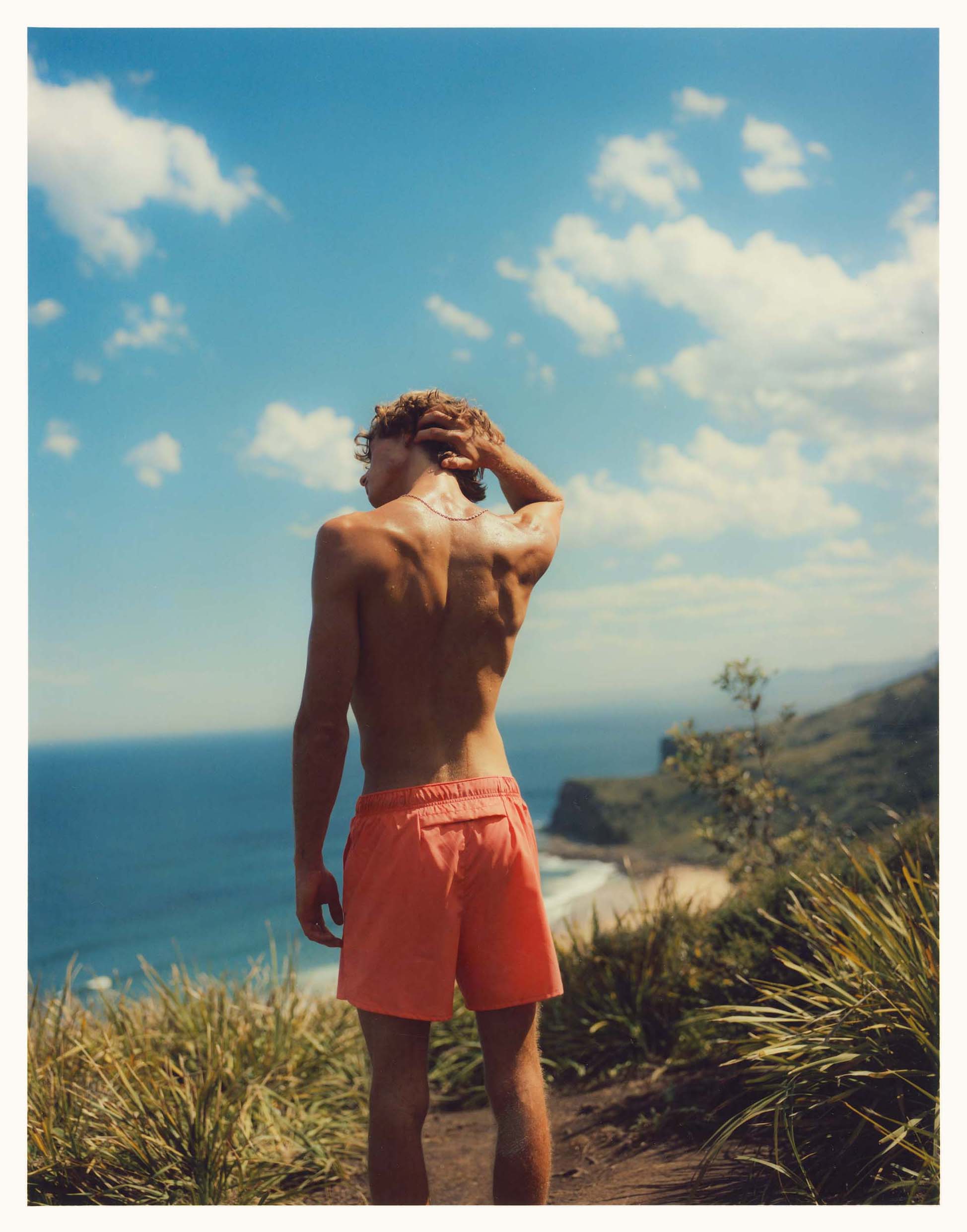 Man with bare torso standing on grassy hilltop overlooking blue ocean and sky with clouds, wearing orange shorts and a necklace.