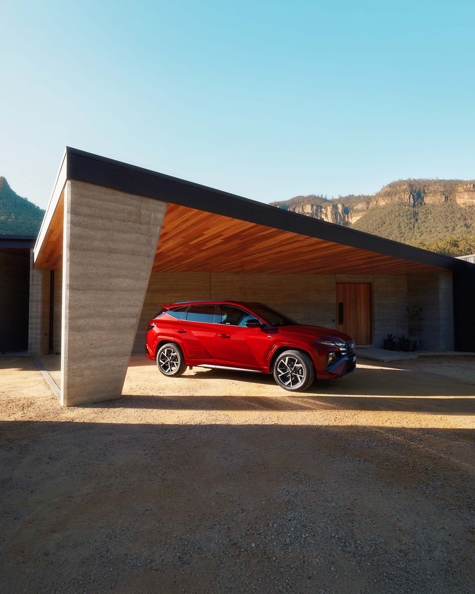 Red SUV parked in front of modern house with flat roof on gravel driveway, with mountains visible in background on sunny day.