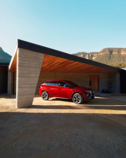 Red SUV parked in front of modern house with flat roof on gravel driveway, with mountains visible in background on sunny day.
