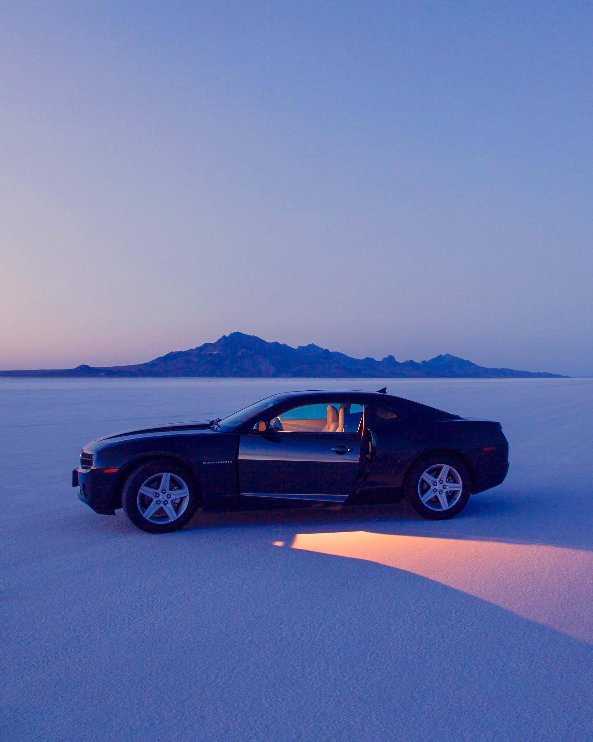 Black car parked on white salt flats with mountains on horizon at dusk. Vehicle has open door, casting a soft glow onto the ground.