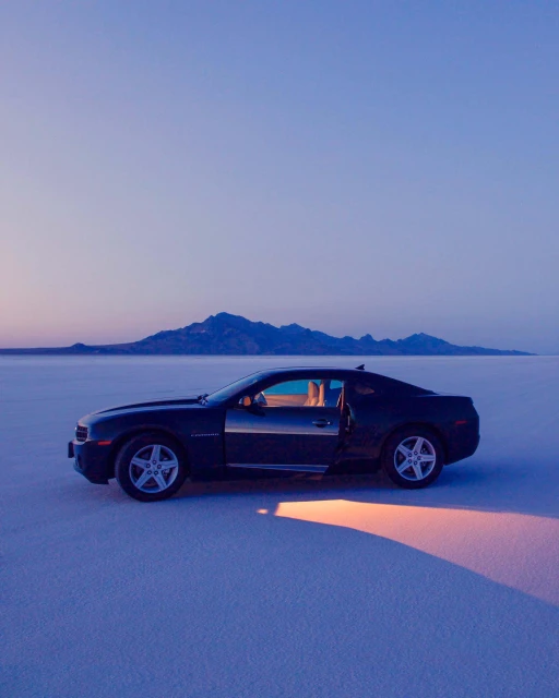 Black car parked on white salt flats with mountains on horizon at dusk. Vehicle has open door, casting a soft glow onto the ground.
