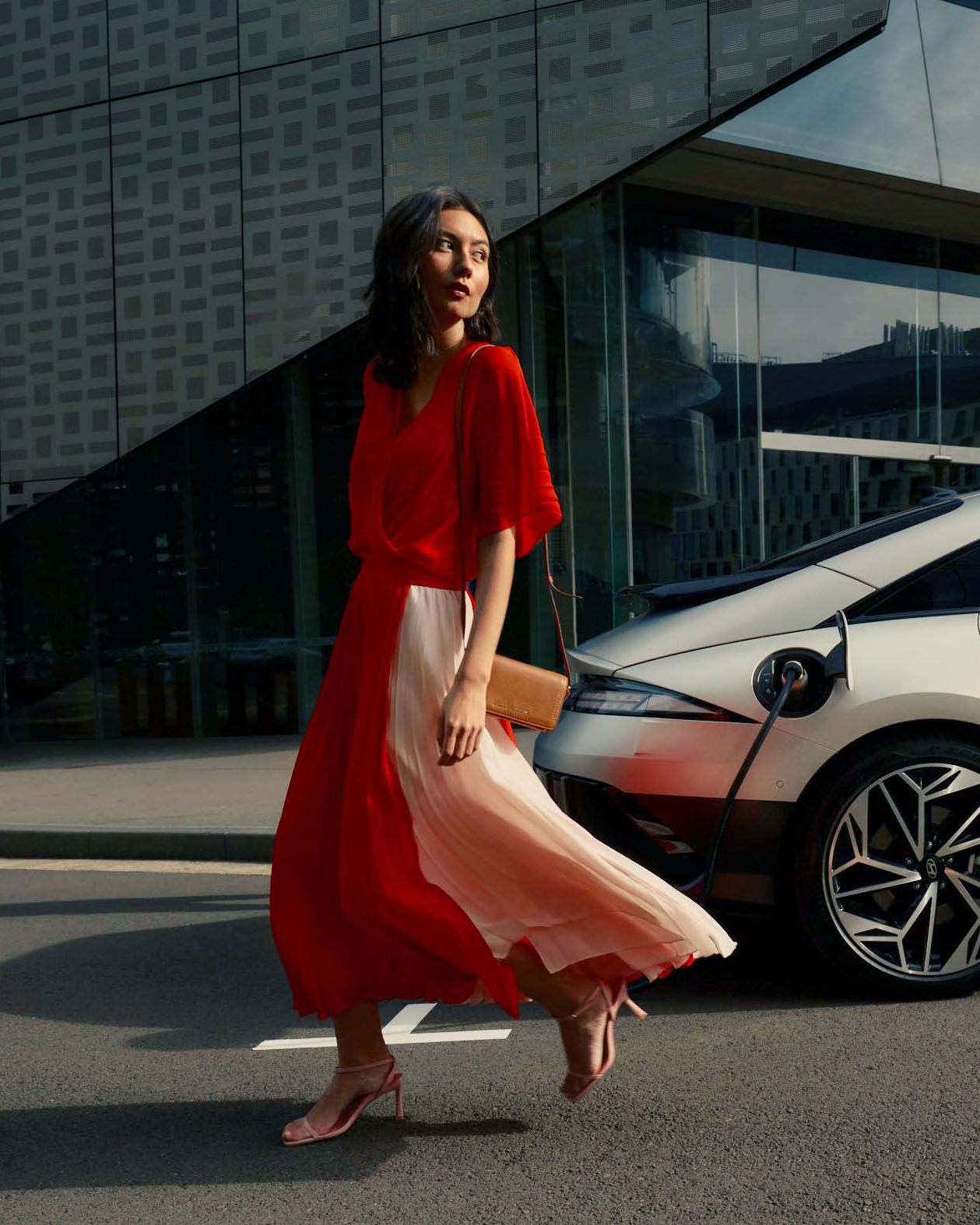 An adult female in a red dress walks in front of a modern building and a white electric car that is plugged into a charger.