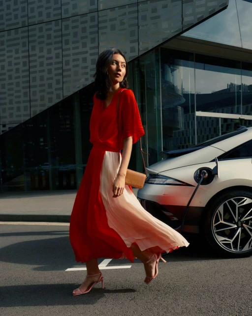 An adult female in a red dress walks in front of a modern building and a white electric car that is plugged into a charger.