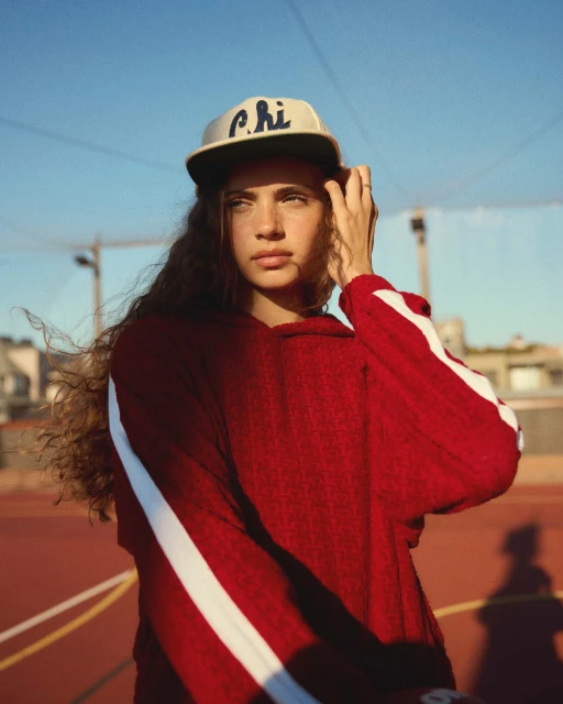 Portrait of a woman with long curly hair wearing a cap and red hoodie with white stripes, standing on a red court under blue sky.