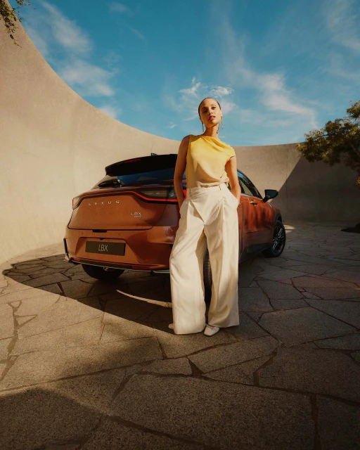 Brown Lexus LBX car parked outdoors with an adult female model posing in front of a curved architectural wall on a sunny day.