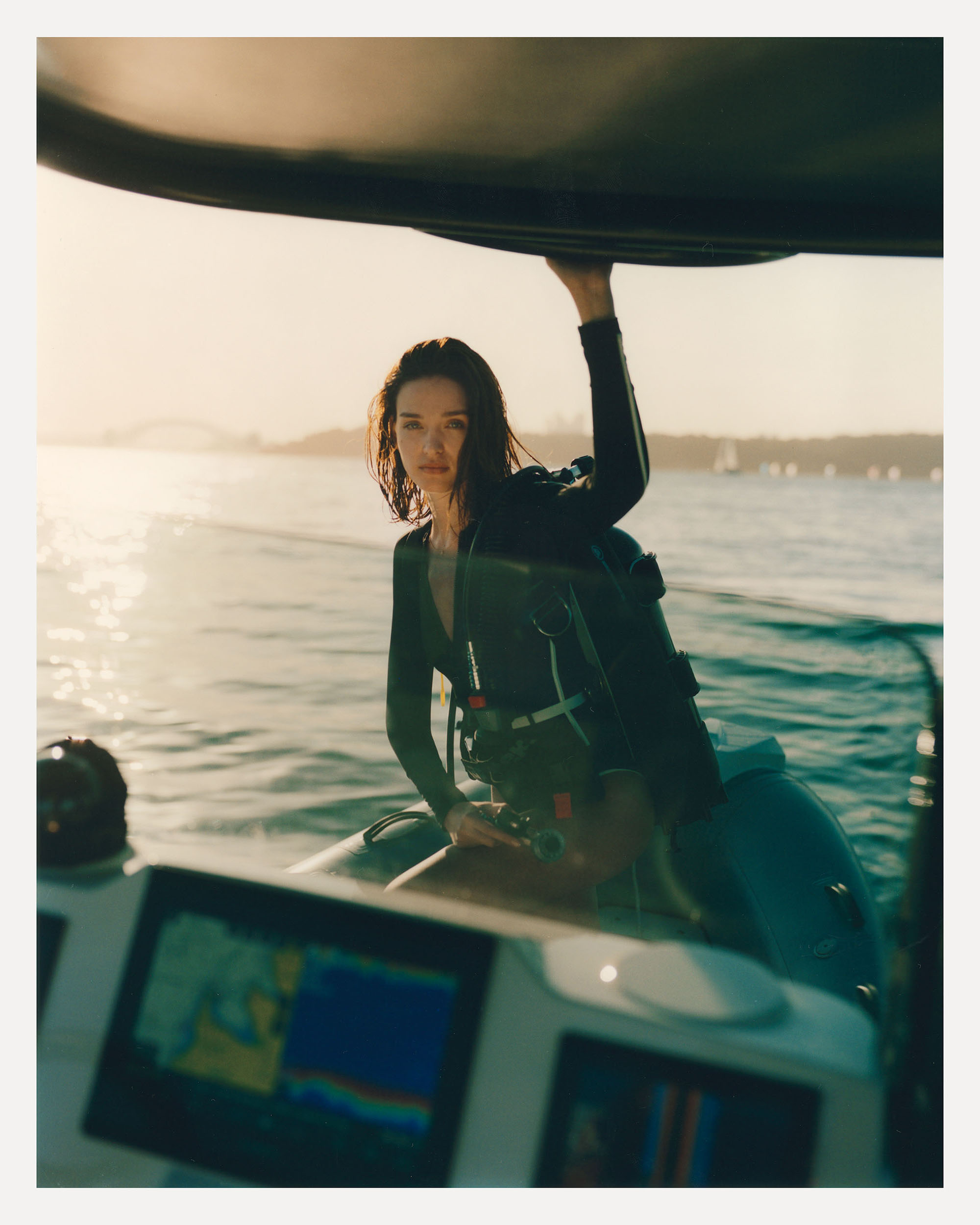 A brunette woman in a wetsuit stands on a boat holding onto the roof, with a blurred seascape and coastline in the background.