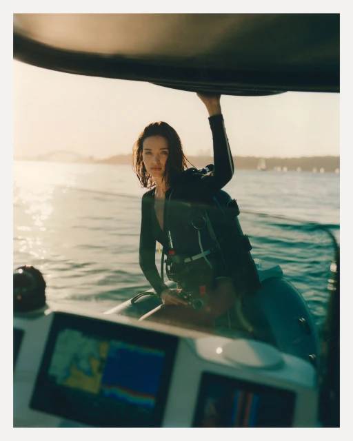 A brunette woman in a wetsuit stands on a boat holding onto the roof, with a blurred seascape and coastline in the background.