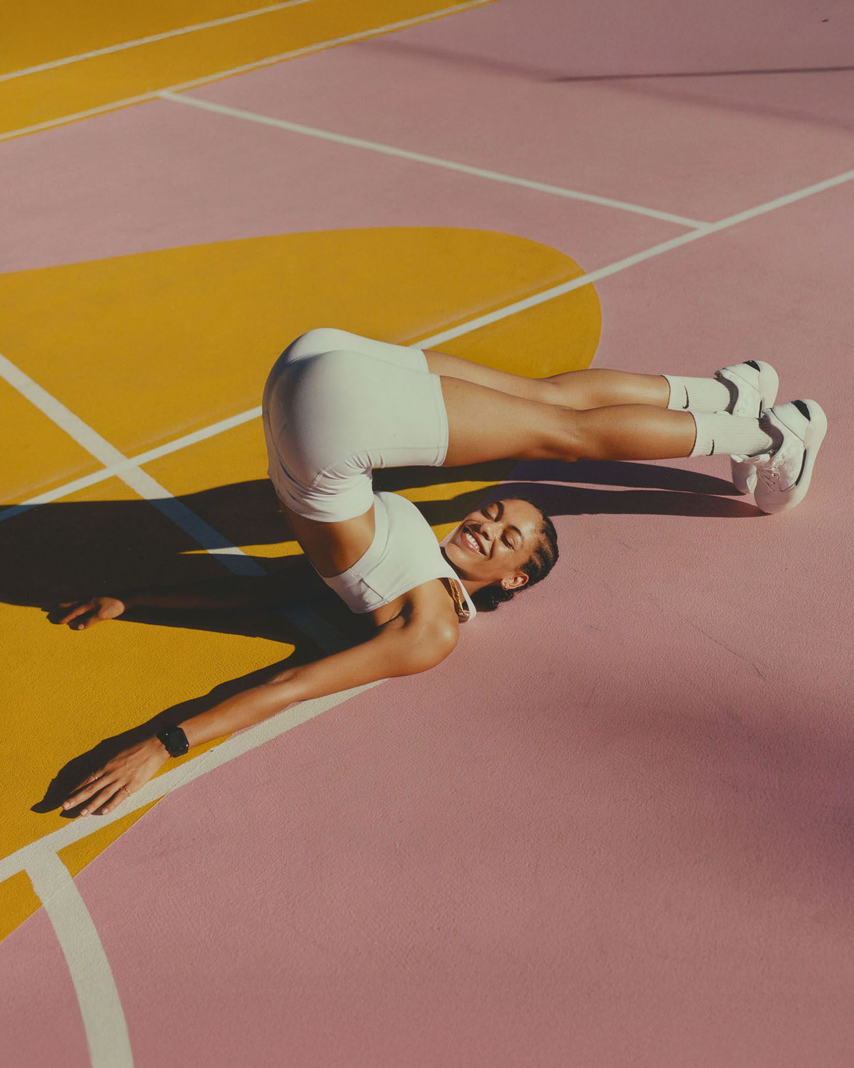 A smiling athletic woman in white activewear does a yoga pose on a pink and gold-painted sports court, captured in eye-level shot.