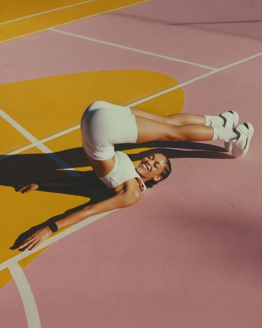 A smiling athletic woman in white activewear does a yoga pose on a pink and gold-painted sports court, captured in eye-level shot.
