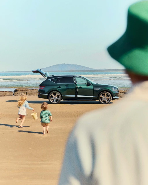 An emerald green Genesis SUV is parked on a sandy beach with two children playing nearby, under a sunny sky near the ocean.