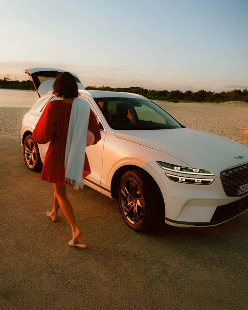Woman in red dress approaches a white Genesis SUV with open trunk on a beach at sunset, carrying a white garment bag.