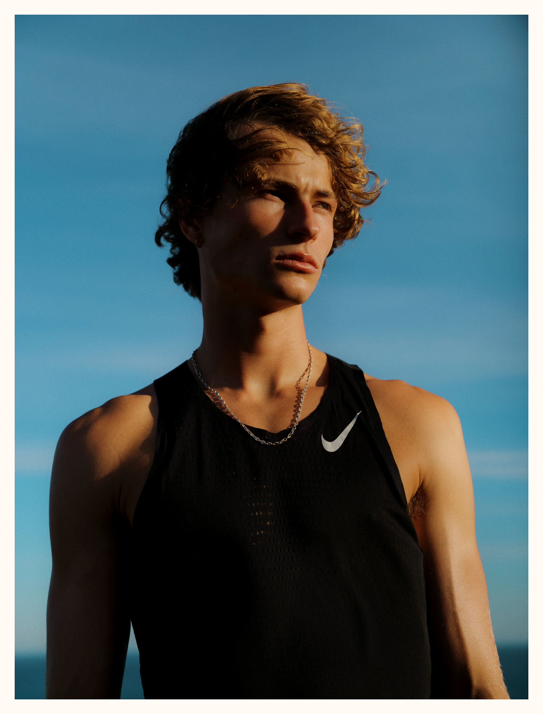 Young man in athletic wear stands outside against a blue sky. He wears a black tank top with a white Nike logo and a silver chain.