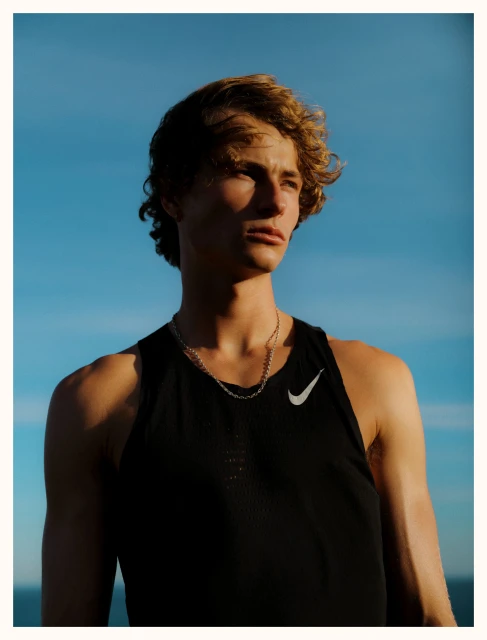 Young man in athletic wear stands outside against a blue sky. He wears a black tank top with a white Nike logo and a silver chain.