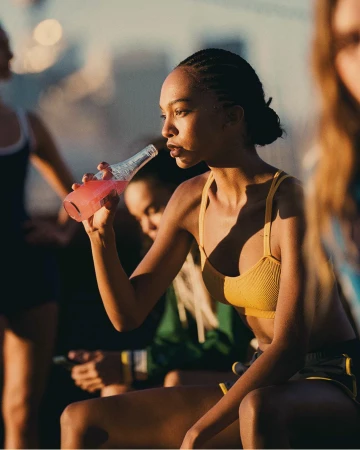 Young Black woman drinking pink beverage from glass bottle wearing yellow sports bra in Sydney by Mark Bramley