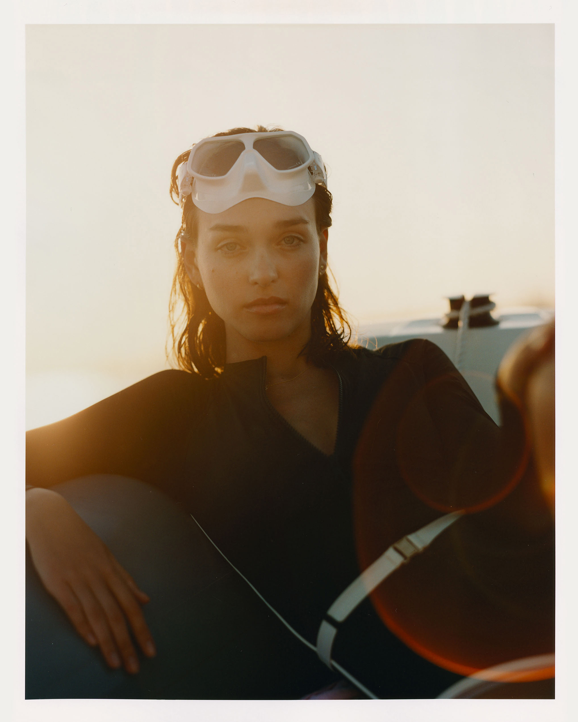 A woman in a wetsuit and goggles poses on a boat against a bright sky, bathed in warm, natural light. Soft focus and a sun-kissed glow.