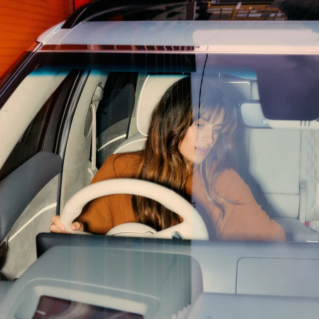 Woman driving modern SUV with luxurious light grey interior and light-colored dashboard, viewed through front windshield in daylight.