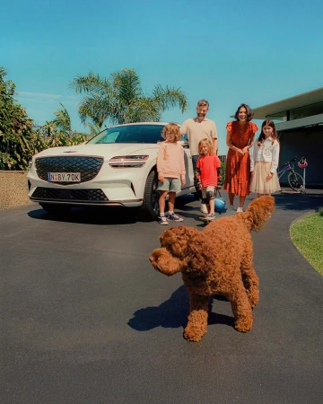 A family of four stands near a white SUV with a dog in the foreground on a driveway near a modern home and landscaping.