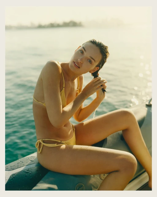 Woman in yellow bikini sits on a boat in the water, running her fingers through her wet hair, with the sea and sky in the background.