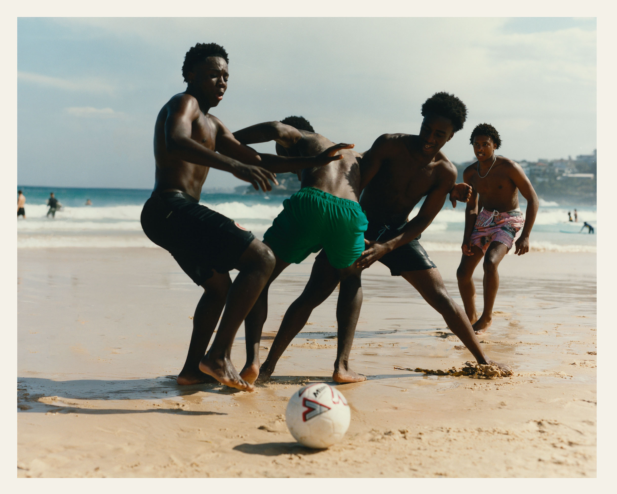 A group of four shirtless young men play soccer with a ball on a sandy beach near the ocean, with other beachgoers and buildings in the distance.
